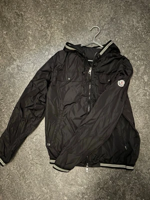 Moncler windbreaker  - Snygg svart vindjacka från Moncler med huva, dragkedja framtill och två bröstfickor med knappar. Jackan har vita och svarta ränder på muddar och krage samt Moncler-logga på ärmen. Tillverkad i lätt, glansigt material som står emot vind.