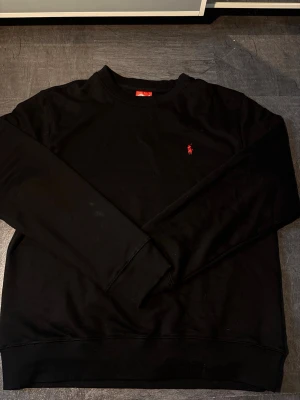 Svart sweatshirt från Ralph Lauren - Svart sweatshirt från Ralph Lauren med klassisk rund hals och den ikoniska röda polospelaren broderad på bröstet. Tröjan har en enkel och stilren design, perfekt för en avslappnad look. Mjuk bomullskvalitet och långa ärmar.