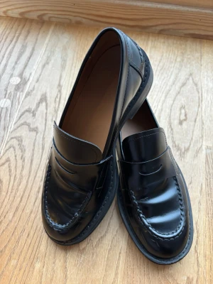 Svarta loafers från Copenhagen Studios - Stilrena svarta loafers i blankt läder från Copenhagen Studios. Klassisk penny loafer-design med rundad tå och diskreta sömmar. Skorna har en låg klack och en slät sula, perfekta för en clean och tidlös look. Nypris 2599kr