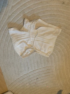 Vita bomullsshorts från Gina Tricot - Säljer ett par vita shorts från Cubus i mjuk bomull med resår i midjan. De har en avslappnad passform och är perfekta för varma dagar. Shortsen är ljusa och har en enkel, stilren look. 