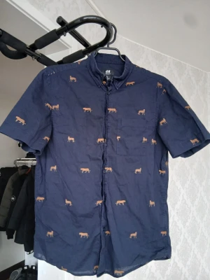 Blå skjorta med leopardmönster H&M - Mörkblå skjorta från H&M med korta ärmar och bröstficka. Skjortan har ett broderat leopardmönster i orange över hela tyget och stängs med knappar framtill. Perfekt för dig som vill sticka ut med en cool och trendig look.