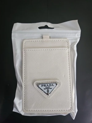 Vit plånbokshållare - Stilren vit plånbokshållare i canvas med klassisk triangelformad logotyp på framsidan. Plånboken har ett fack på framsidan och ett transparent fack på baksidan, samt en matchande vit rem med metallspänne. Perfekt för kort och ID.