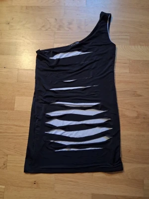 Svart oneshoulder topp - Säljer en svart topp med oneshoulder och flera slitsar framtill för en edgy look. Toppen har tight passform och är tillverkad i ett stretchigt material som sitter snyggt på kroppen.