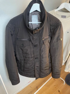 Mörkbrun field jacket från J.Lindeberg  - Snygg field jacket från J.Lindeberg i en fin mörkbrun färg. En knapp saknas som visas på bild 5 annars är jackan i helt nyskick. Hör gärna av er vid intresse!