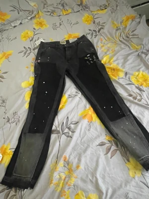 Svarta jeans med färgstänk och patchar - Unika svarta jeans från Pull&Bear med coola färgstänk och patchar på benen. Jeansen har raka ben, flera fickor och detaljer som ger en streetwear vibe. Materialet är denim och färgen går från svart till grått nedtill. Perfekta för dig som gillar en edgy stil.