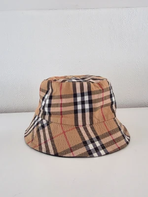 Beige Burberry bucket hat med rutigt mönster - Snygg bucket hat i klassiskt beige rutigt mönster med svarta, vita och röda linjer. Hatten har en mjuk form och är tillverkad i bomullstyg. Perfekt accessoar för att lyfta din stil med en trendig touch.