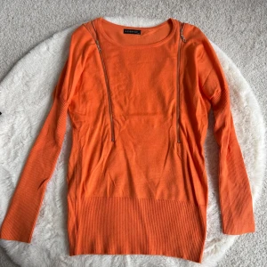 Orange stickad tröja med dragkedjor (XS/S) - Snygg orange stickad tröja med långa ärmar och två dekorativa dragkedjor framtill. Tröjan har bred ribbstickad kant nedtill och vid ärmsluten. Perfekt för dig som vill ha en färgklick i garderoben.