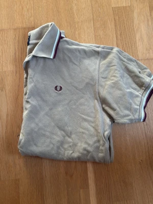 Beige pikétröja från Fred Perry - Klassisk beige pikétröja från Fred Perry med korta ärmar och krage med vita och vinröda ränder. Tröjan har den ikoniska Fred Perry-loggan broderad på bröstet och är tillverkad i bomull. Perfekt för en avslappnad och stilren look.