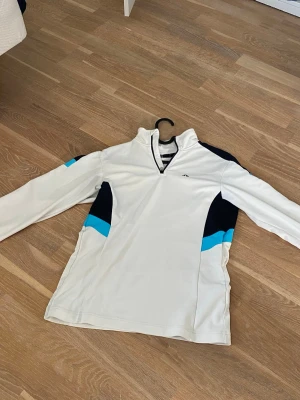 Vit sporttröja från J.Lindeberg - Snygg vit långärmad sporttröja från J.Lindeberg med halv dragkedja. Tröjan har marinblå och ljusblå detaljer på sidorna och ärmarna. Tillverkad i ett stretchigt funktionsmaterial som passar perfekt till träning eller golf.