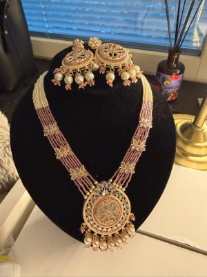 Indiskt halsband med pärlor och örhängen - Säljer ett vackert indiskt halsband med matchande örhängen. Halsbandet har flera rader av vita och rosa pärlor, dekorerat med guldfärgade blommor och en stor rund medaljong med detaljer. Örhängena är stora, runda och prydda med pärlor och guldfärgade detaljer.
