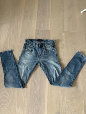 Blå jeans från Replay - Säljer ett par blå slim fit jeans från Replay med klassisk femficksdesign och slitningar på låren. Jeansen har en snygg tvättad look, normalhög midja och Replays ikoniska läderpatch bak i midjan. Perfekta för en avslappnad och trendig stil. Pris går att diskutera. Vid intresse är det bara att höra av sig 