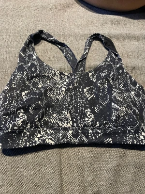  sport-bh med mönster - Säljer en svart sport-bh med vitt ormliknande mönster och korsade axelband i ryggen. Bh:n har bred resår under bysten och är tillverkad i ett stretchigt material som passar perfekt för träning eller en aktiv vardag.