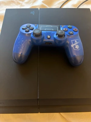 PlayStation 4 med blå specialkontroll - Säljer en svart PlayStation 4-konsol från Sony tillsammans med en blå specialutgåva av DualShock 4-kontrollen med unikt mönster och PlayStation-logga. Konsolen har en stilren, rektangulär design i matt plast och passar perfekt för gamingkvällar.