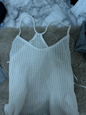Vit stickad linne med smala band - Säljer ett vitt stickat linne med smala axelband och v-ringning. Toppen har en luftig och transparent struktur som ger en avslappnad vibe. Perfekt för lager på lager och enkel att styla till olika looks.