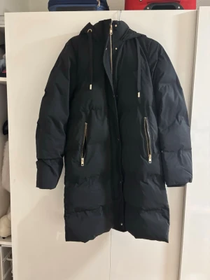 Svart lång dunjacka med huva - Säljer en svart lång dunjacka med huva och dragsko. Jackan har två stora fickor med guldfärgade dragkedjor och en rak passform. Perfekt för kalla vinterdagar och riktigt varm tack vare det tjocka dunfodret.