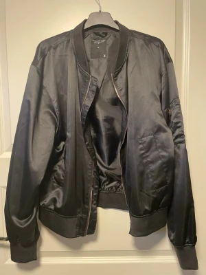 Svart tunn jacka   - Snygg svart tunn jacka  med glansig finish. Klassisk modell med ribbade muddar, dragkedja och fickor på sidorna samt en ficka med dragkedja på ärmen. Perfekt för en trendig streetwear-look.