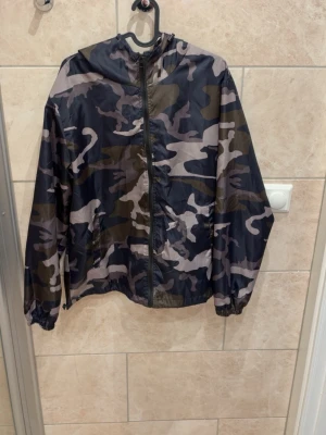 Camouflage vindjacka från Lindfors - Snygg vindjacka från Lindfors med klassiskt camouflage-mönster i grönt, grått och svart. Jackan har huva, dragkedja framtill och elastiska muddar vid ärmsluten. Perfekt för dig som vill ha en lätt och stilren jacka till vardags.