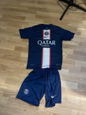 PSG Neymar Jr matchställ blå Nike  - Snyggt PSG matchställ i marinblått med Neymar Jr och nummer 10 på ryggen. Tröjan har vita och röda detaljer, Qatar Airways sponsortryck och PSG-logga. Shortsen har elastisk midja, PSG-märke och Nike-logga. Perfekt för fotbollsträning eller att bära som supporter.