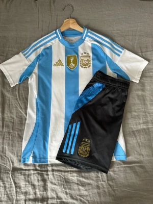 Argentina Landslagskit - Argentina Landslagskit (Tröja + Shorts) – Stl 164 Säljer ett komplett och jättefint set med Argentinas hemmatröja och tillhörande shorts från Adidas. Kitet är i mycket gott skick och har inga fläckar, hål eller slitningar. • Storlek: Junior 164 (13–14 år). Som passar XS/S. • Skick: Sparsamt använt, ser nästan nytt ut. • Material: Adidas officiella funktionsmaterial som andas bra. • (Nypris ca 1 000 kr). 