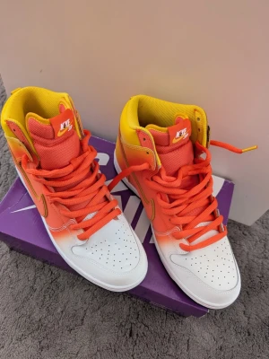Nike SB Dunk High Begränsad upplaga  orange/gul Storlek 45,5 - Nike SB Dunk High sneakers i en färgstark mix av orange, gult och vitt. Skorna har en hög siluett, orange snörning och swoosh, samt vit tåbox med perforeringar. Ovandelen är i läder och mesh för både stil och komfort. Perfekta för dig som vill sticka ut.