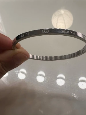 Silverfärgat armband från Gucci - Snyggt och stilrent armband från Gucci i silverfärgad metall. Armbandet är stelt och har ingraverad Gucci-logga samt dubbla G-symboler runt om. Perfekt accessoar för dig som gillar exklusiva detaljer och enkel design. Rostfritt 