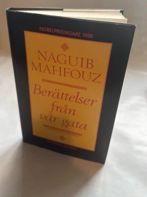 Berättelser från vår gata - Mahfouz - En inbunden bok med svart och gul omslag, skriven av Naguib Mahfouz, Nobelpristagare 1988. Titeln 'Berättelser från vår gata' står i röd text på framsidan. Perfekt för dig som gillar klassisk skönlitteratur.