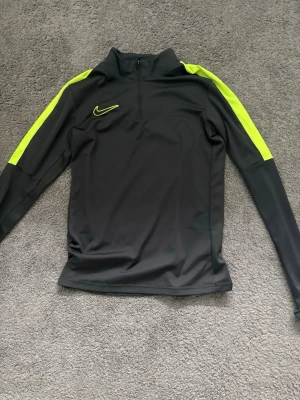 Nike svart träningsöverdel med neon - Nike långärmad träningsöverdel i svart med neonfärgade detaljer längs axlar och ärmar. Halvlång dragkedja framtill och sportig passform. Perfekt för träning eller uppvärmning. Tillverkad i ett lätt och stretchigt funktionsmaterial.