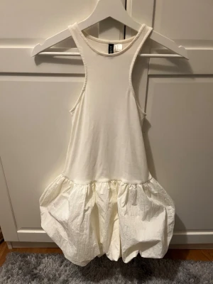 Vit ärmlös klänning med ballongkjol - Säljer en vit ärmlös klänning med ribbad överdel och ballongformad kjol. Klänningen har rund halsringning och breda axelband. Materialet upptill är stretchigt medan kjolen är i ett krispigare tyg. Perfekt för en trendig och avslappnad look.