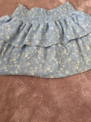 Blå blommig volangkjol från Gina Tricot - Supersöt blå kortkjol från Gina Tricot med vita små blommor och volangdetalj. Kjolen har bred smock i midjan för skön passform och är perfekt för dig som gillar en feminin och trendig stil.