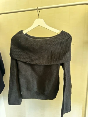 Svart off shoulder stickad tröja - Mysig svart stickad tröja med bred ribbad off shoulder-krage och långa ärmar. Tröjan har en avslappnad passform och ribbade muddar vid ärmslut och nederkant. Perfekt för dig som gillar en trendig och bekväm stil.