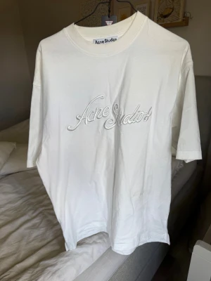 Vit Acne Studios logo t-shirt - Säljer en vit t-shirt från Acne Studios med broderad logga framtill. T-shirten har avslappnad passform, rund halsringning och korta ärmar. Tillverkad i mjuk bomull för en clean och stilren look.