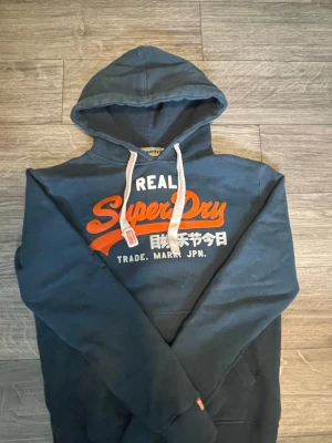 Mörkblå Superdry hoodie med tryck - Mörkblå hoodie från Superdry med stor orange och vit logga på bröstet samt japansk text. Klassisk huva med vita dragsnören och mjukt, borstat innerfoder. Perfekt för dig som gillar streetwear och vill ha en snygg och bekväm hoodie.