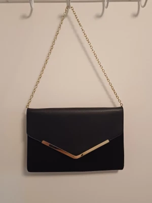 Svart kuvertväska med guldkedja - Stilren svart kuvertväska med guldfärgad kedja och snyggt gulddetaljerat V på locket. Väskan har en klassisk rektangulär form och är tillverkad i ett slätt material som ger en elegant look. Perfekt att bära över axeln med kedjan. Skriv om vi har frågor, priset går att diskutera ❤️