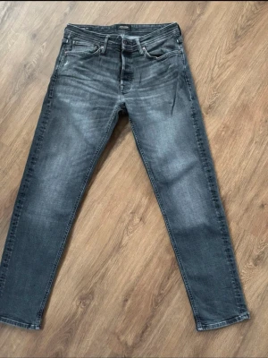 Jack& Jones jeans -Taperd Mike - 32/32 Snygga grå jeans med tvättad och lätt slitad look. Klassisk femficksmodell med dragkedja och knapp i midjan. Jeansen har raka ben och är perfekta för en avslappnad stil. Materialet är troligtvis bomull med lite stretch för extra komfort.