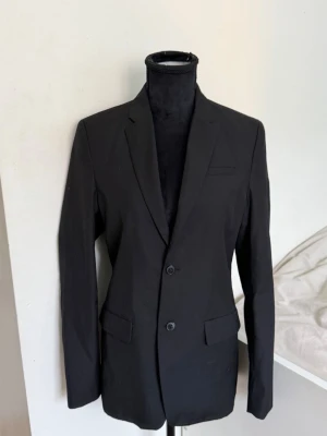 Svart klassisk kavaj med två knappar - Stilren svart kavaj med två knappar framtill och klassiska slag. Kavajen har två fickor med lock och en bröstficka. Passar perfekt till både skjorta och t-shirt för en snygg look. Materialet ser ut att vara en blandning av polyester och bomull.