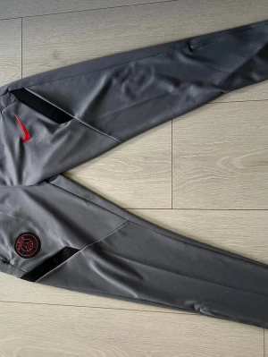 Grå PSG träningsbyxor från Nike - Snygga grå träningsbyxor från Nike med PSG-logga och röd Nike Swoosh på ena benet. Byxorna har smal passform, dragkedjor vid bensluten och sidofickor. Perfekta för fotbollsträning eller chill. Tillverkade i ett mjukt och stretchigt material.