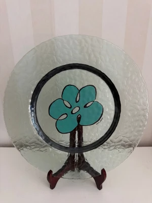 Annika Scheller glastallrik med blommönster - Unikt rund glasfat med hamrad yta och ett turkost blommotiv i mitten, omgiven av en svart cirkel. Fatet har en konstnärlig känsla och designad av Annika Scheller.