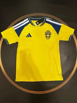 Sverige matchtröja Adidas gul/blå - Gul matchtröja från Adidas med blå detaljer och vita ränder på axlarna. Tröjan har Sveriges landslagsemblem på bröstet och texten 'Sverige' på ryggen. Tillverkad i lätt och ventilerande material, perfekt för fotboll eller träning.