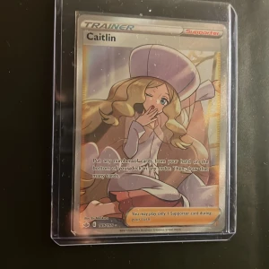 Pokémonkort Caitlin Trainer Supporter - Säljer ett Pokémonkort med Caitlin Trainer Supporter-motiv. Kortet har en färgglad illustration med guldfärgade detaljer och text på engelska. Kortet ligger i ett genomskinligt plastfodral för extra skydd. Perfekt för samlare eller Pokémonfans.