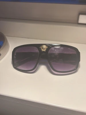 Svarta solglasögon med gulddetalj - Snygga svarta solglasögon från  med breda skalmar och en ikonisk guldfärgad Medusa-detalj på sidan. Glasen är mörka och bågarna har en klassisk, rektangulär form. Perfekta för dig som vill sticka ut med en lyxig accessoar.