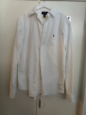 Vit skjorta från Polo Ralph Lauren - Klassisk vit skjorta från Polo Ralph Lauren med broderad logga på bröstet. Skjortan har lång ärm, knappar framtill och en tidlös design som passar till det mesta. Tillverkad i bomull för en skön känsla.