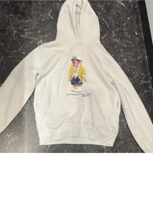 Vit hoodie Polo Ralph Lauren björn - Vit hoodie från Polo Ralph Lauren med den ikoniska Polo Bear-trycket på bröstet. Björnen har på sig en gul jacka, randig tröja och keps. Hoodien har dragsko i huvan och ribbade muddar. Perfekt för dig som gillar streetwear och klassiska prints.