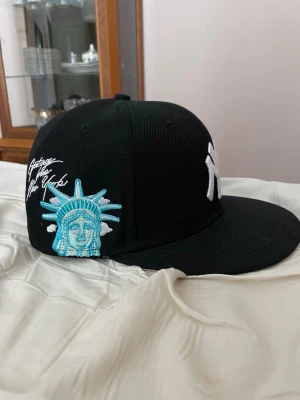 Svart New York Yankees keps - Säljer en svart New York Yankees keps från New Era med vit broderad NY-logga framtill och broderad frihetsgudinna i blått och vitt på sidan. Kepsen har platt skärm och klassisk snapback-stil. Perfekt för dig som gillar streetwear och sportig look.