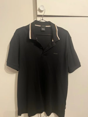 Svart polotopp från BOSS slim fit oanvänd  - Snygg svart polotopp från BOSS i slim fit-modell. Kragen har vita och beige detaljer som ger en clean kontrast. Klassisk pikélook med korta ärmar och diskret BOSS-logga på bröstet. Perfekt till jeans eller chinos för en stilren vibe.