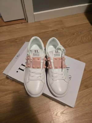 Valentino Rockstud Untitled sneakers - Säljer ett par vita Valentino Rockstud Untitled sneakers med ljusrosa band och ikoniska nitar över snörningen. Skorna har en clean och modern design med tydlig logga på plösen. Tillverkade i slätt läder och passar perfekt för dig som gillar exklusiva detaljer.