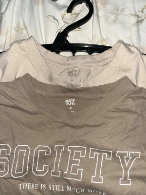  t-shirt  - Snygg beige t-shirt från 157 med trycket 'SOCIETY' och texten 'THERE IS STILL MUCH MORE' på bröstet. Klassisk rund halsringning och korta ärmar. Tillverkad i mjuk bomull som känns skön mot huden. Perfekt för en avslappnad och trendig stil.