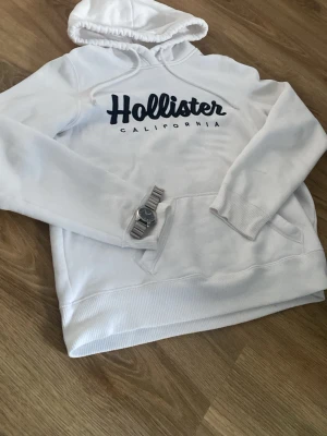 Vit hoodie från Hollister - Snygg vit hoodie från Hollister med stor svart logga och texten 'California' på bröstet. Hoodien har huva med dragsko och en klassisk magficka. Tillverkad i mjukt material som känns skönt mot huden. Perfekt för en avslappnad och trendig look. 