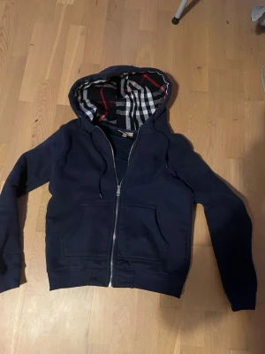 Marinblå hoodie från Burberry - Snygg marinblå hoodie från Burberry med dragkedja framtill och klassiskt rutigt foder i huvan. Hoodien har långa ärmar, fickor framtill och justerbar huva med snören. Perfekt för dig som vill ha en stilren och exklusiv look.