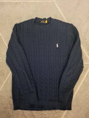 Mörkblå kabelstickad tröja Polo Ralph Lauren - Den är lite knoppig.Kabelstickad mörkblå tröja från Polo Ralph Lauren med rund halsringning och den klassiska broderade loggan på bröstet. Tröjan har långa ärmar och ribbade muddar vid ärmslut och nederkant. Perfekt för dig som gillar stilren och tidlös design.