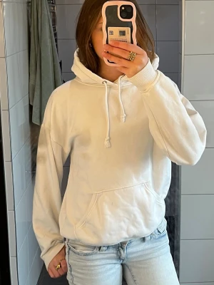 Vit hoodie med ficka framtill - Klassisk vit hoodie med huva och dragsnören, samt en stor känguruficka framtill. Tillverkad i mjukt material och har ribbade muddar vid ärmslut och nederkant. Perfekt för en avslappnad och stilren look.